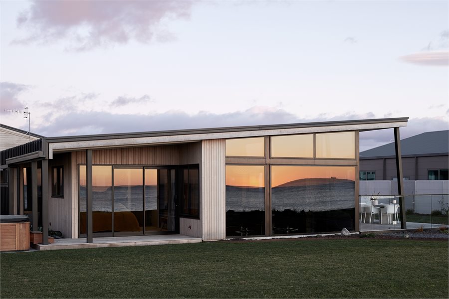 Big picture windows with raked windows above take in the view from the lounge floor to ceiling. The home features redwood timber stained with Dryden WoodOil and is complemented by Matt Black powdercoated aluminium joinery.